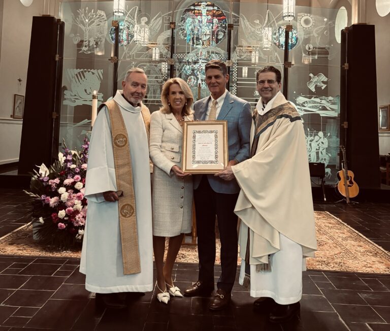 Four people stand in a church holding a framed certificate. Two priests in white liturgical vestments flank a woman in a cream suit and a man in a light blue jacket. They are positioned in front of a modern etched-glass altar screen.