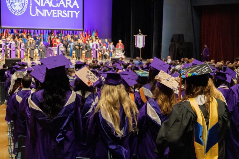 A high-angle view from behind a crowd of Niagara University graduates wearing purple caps and gowns during a commencement ceremony at Artpark.