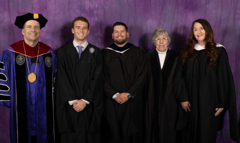 Five people standing against a purple backdrop. All are dressed professionally and some in graduation garb.