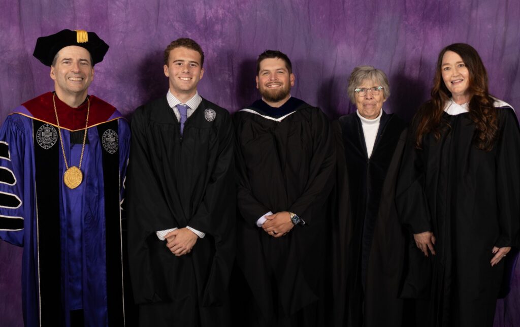 Five people standing against a purple backdrop. All are dressed professionally and some in graduation garb.