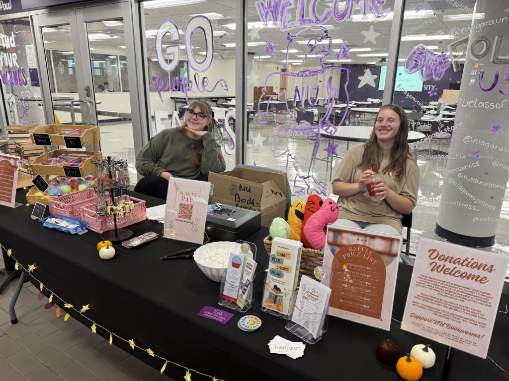 Two smiling students sit behind a black-draped table at the NU Bookworms Fall Book Fair.