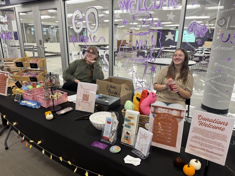 Two smiling students sit behind a black-draped table at the NU Bookworms Fall Book Fair.