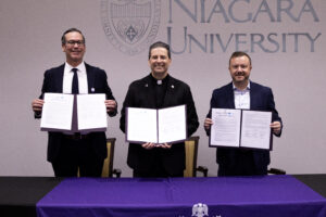 Stanley Wojton, the Rev. James J. Maher, C.M., and David Rust hold up a signed memorandum of understanding in front of a Niagara University backdrop.