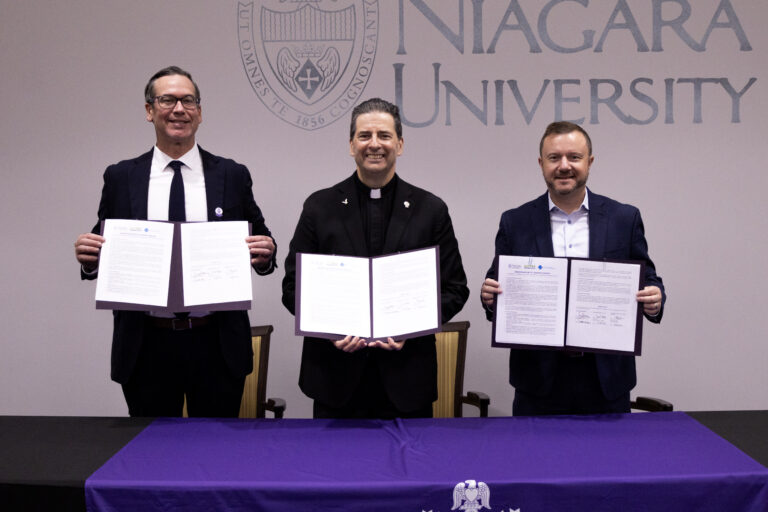 Stanley Wojton, the Rev. James J. Maher, C.M., and David Rust hold up a signed memorandum of understanding in front of a Niagara University backdrop.